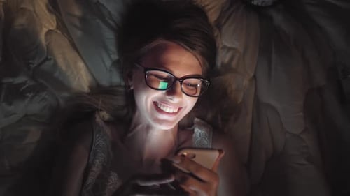 Girl Watching Comedy Movie at Night in Bed. Tablet Smart Phone or Laptop Glowing on Womans Face