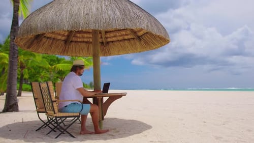 Man Working on Laptop Under Umbrella on Beach