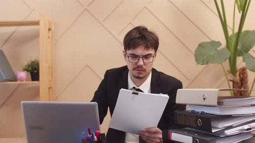 Male manager is working intently at an office desk, holding documents in hand and looking at laptop