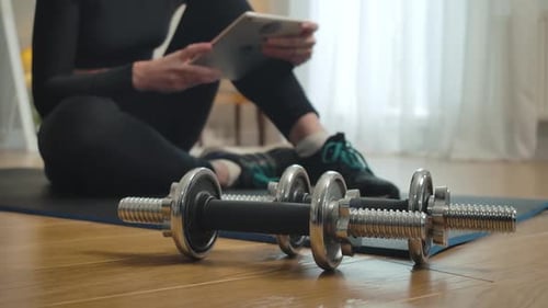 Close-up of Dumbbells with Blurred Slim Sportswoman Surfing Internet on Tablet at the Background