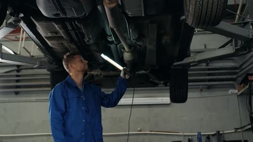Car Mechanic Examining Car Suspension of Lifted Automobile at Repair Service Station with Light