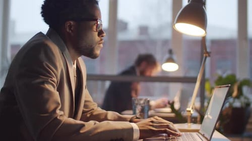 Afro-American Male Office Worker Using Laptop at Desk in the Evening