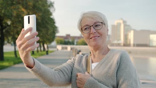 Beautiful Senior Lady Taking Selfie with Smart Phone Camera Posing at River Embankment on Summer Day