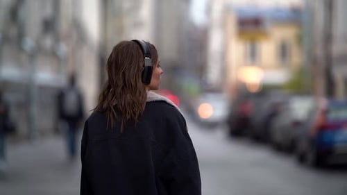 Woman Is Walking in City at Cold Autumn Day, Listening To Music By Headphones, Rear View in Movement