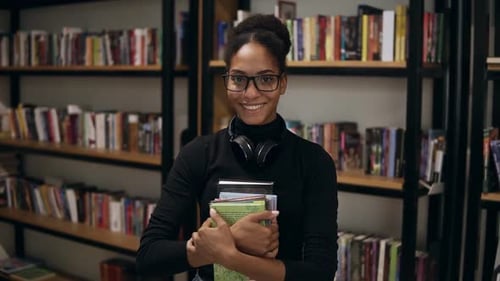 Young African Woman Standing in Library Holding Books Smiling Cheerful Looking Camera Surprised
