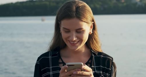 Close Up Portrait of Young Attractive Woman Smiling and Looking at Smartphone Near Beach at Sunset