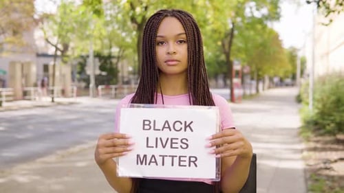 A Young Black Woman Shows a Black Lives Matter Sign To the Camera in the Street in an Urban Area