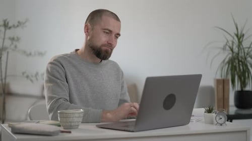 Sleepy male freelancer with beard in a gray jacket works at a laptop at home.