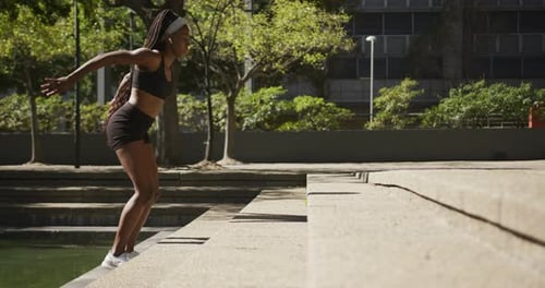 African american woman wearing wireless earphones exercising outdoors jumping on stairs in the city