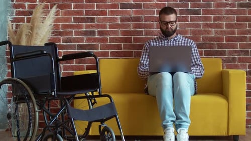 Man Works on Laptop Beside Wheelchair at Home