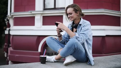A Fashionable Girl in Glasses Sits on the Street on a Parapet Concentrates on Her Mobile Phone