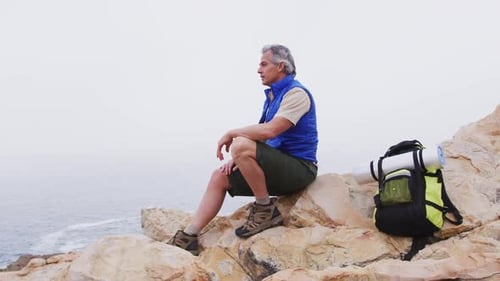 Senior hiker man with backpack sitting on the rocks in the mountains and watching the sea shore.
