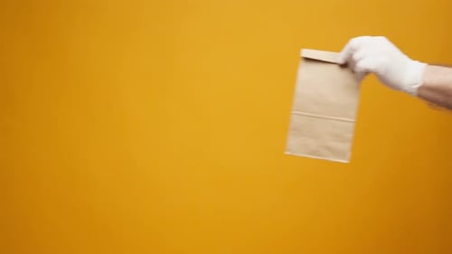 Courier in Gloves Passing Craft Shopping Bag with Delivery Against Yellow Background