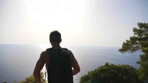 Hiker with Backpack Reaching Up Edge of Mountain and Raising His Hands Admiring Scenic Seascape. Man
