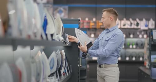 A Young Handsome Man in a Shirt Chooses an Iron in a Consumer Electronics Store. Bachelor