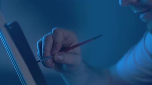 Busy Man Artist with a Brush in His Hand Picks Up an Oil Painting in a Cozy Studio