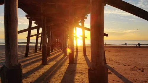 Drone flying fast under Newport Beach pier at sunset on the ocean with people walking the California