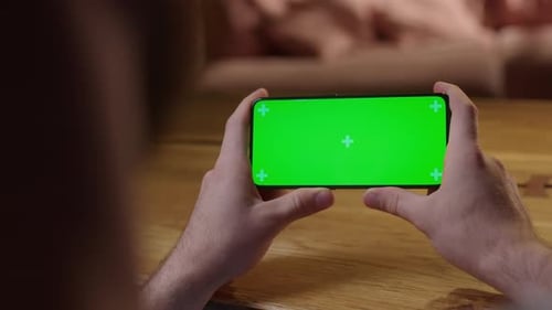 Handheld Camera Back View of Young Man at Home Sitting on a Wood Desk Using With Green Mockup Screen