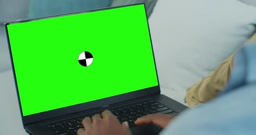 African Man Lying on Sofa at Home Uses Laptop with Green Mockup Screen