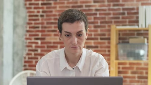 Shorthaired Adult Woman in White Shirt Works Behind a Laptop and Takes Sip of Coffee From Paper Cup
