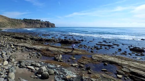 Side view tracking shot of a young man running on a rocky ocean beach shoreline.