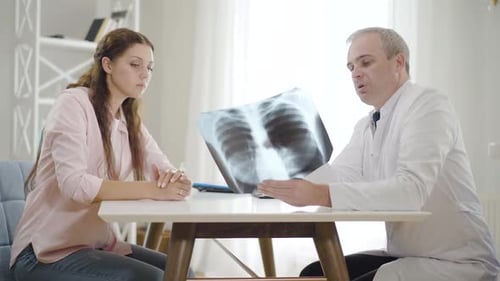 Professional Physician Explaining Lungs X-ray To Young Woman in Hospital, Side View Portrait of Mid