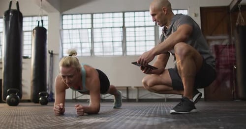 Fit caucasian woman performing plank exercise with male trainer holding digital tablet at the gym