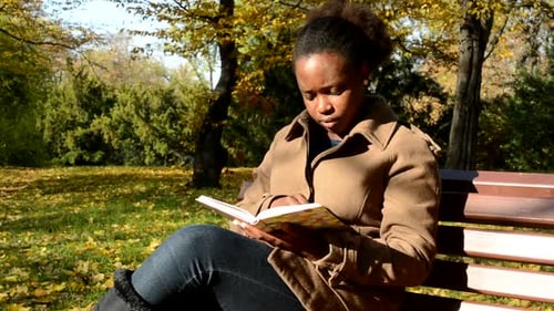 Joven y hermosa niña africana tranquila se sienta en un banco en el bosque, examina un libro y observa el paisaje