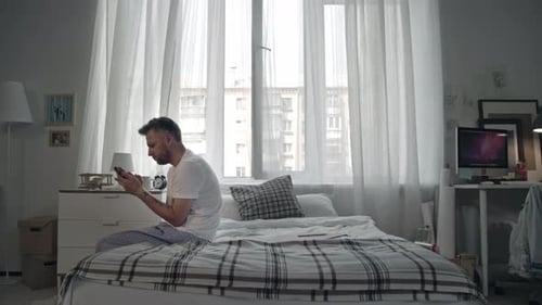 Man Sits on Bed Using Smartphone in Bedroom