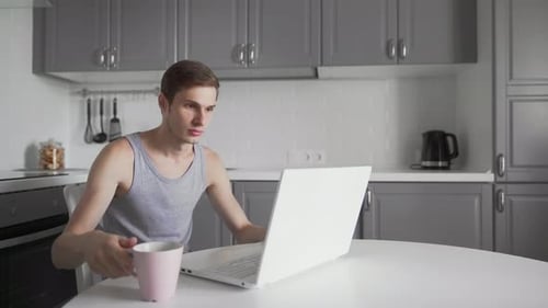Young Adult Using Laptop in Kitchen, Drinking Beverage