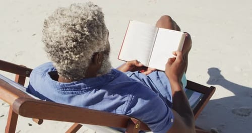 Senior african american man reading and lying on sunbed on sunny beach