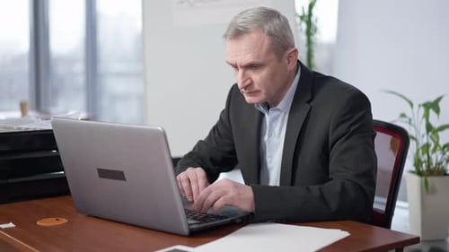 Concentrated Senior Caucasian Man Sitting in Office Working Online on Laptop
