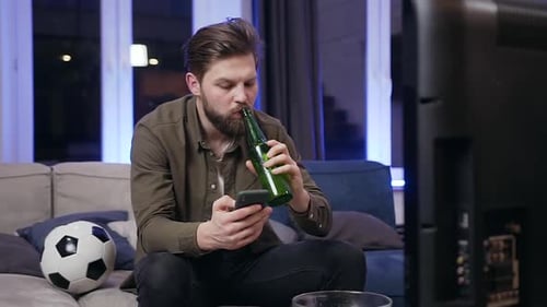 Young Bearded Man which Drinking Beer while Browsing His Phone in Front of TV
