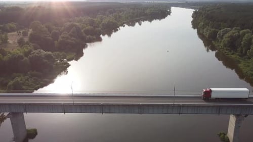 Semi Truck with Cargo Trailer Passing Highway Bridge over the river, in the rays of the setting sun.