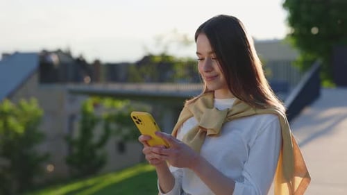 Portrait of Young Woman with Brown Hair Standing on Street and Typing Messages on Smartphone