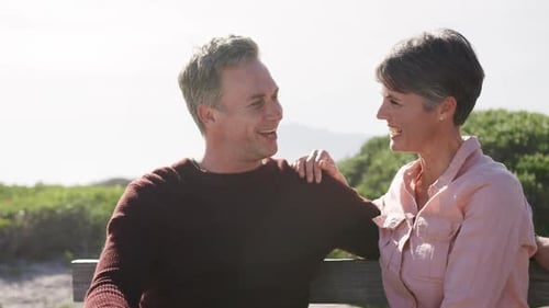 Caucasian couple enjoying free time by sea on sunny day sitting on bench and talking