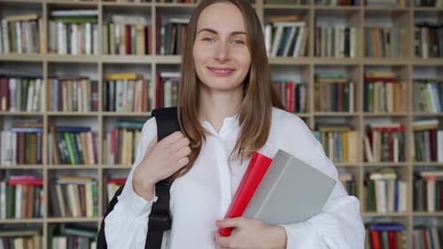 Young Smiling Woman Stands with Books and a Backpack in the Library