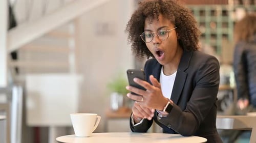 Upset African Businesswoman Reacting To Loss on Smartphone at Cafe