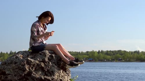 A Young Woman Sits on a Stone By the River with a Phone in Her Hands. The Girl Is Texting in the