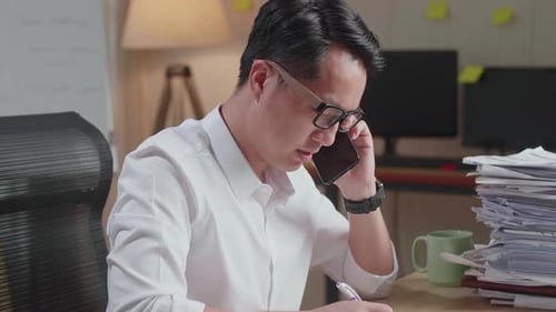 Close Up Of Asian Man Talking On Phone And Taking Note While Working Hard With Documents At Office
