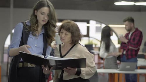 Young and Mature Pretty Women Discussing Project Standing in the Office with Big Paper Folder