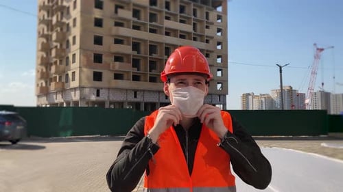 Male Construction Worker in Overalls Putting on Medical Mask on Face on Construction Area.
