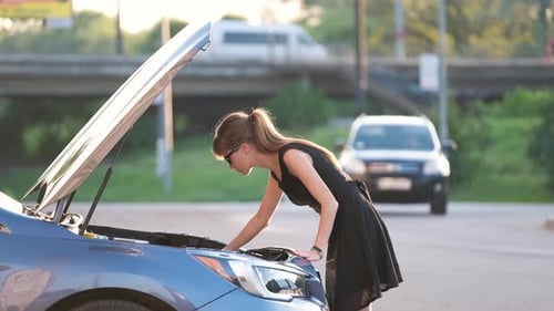 Puzzled Female Driver Standing on a City Street Near Her Car with Popped Up Hood Looking at Broken
