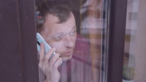 Young Man Outside the Window Looking at the Street and Talking on a Mobile Phone