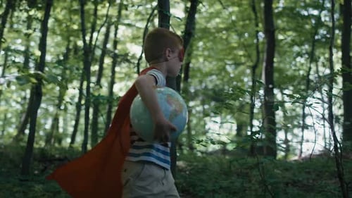 Ginger caucasian boy with superhero costume holding globe and walking in the forest. Shot with RED h