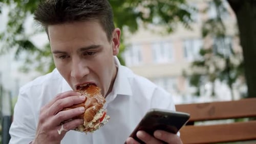 Man Eats Burger and Looks at Phone Outside