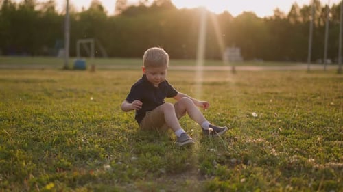 Little Boy Kicks Legs Sitting on Lush Grass on Playground
