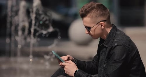 Young Man in Sunglasses Using Phone Sitting Near Fountain