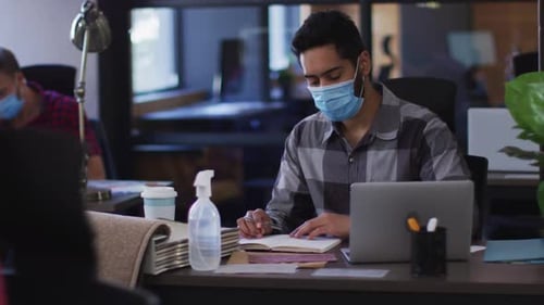 Middle eastern man wearing face mask taking notes and using laptop at modern office