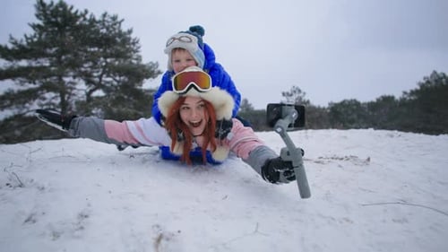 Fun Winter Holidays Joyful Woman with Her Son Slides Down a Snowy Hill on a Sled with a Mobile Phone